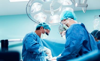 three people in blue gowns, surgical masks and gloves stand under a bright light looking down